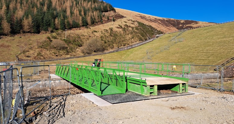 A large, green temporary bridge structure for construction or maintenance purposes, set up in a rural landscape with a backdrop of tree-lined hills under a clear blue sky.