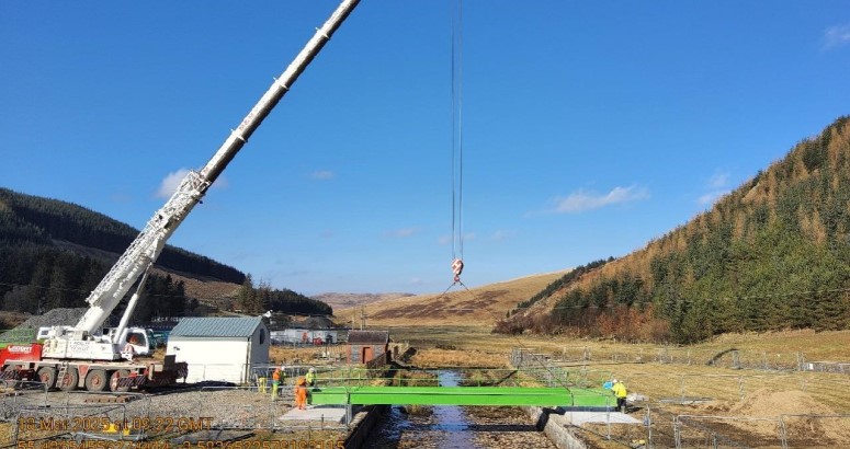 A large crane installing a green bridge in a rural landscape under a clear blue sky.