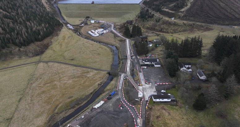 Aerial view of a winding road through a rural landscape with visible buildings and vehicles, surrounded by hills and a river.