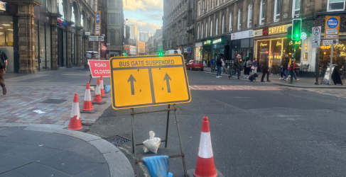 Union Street Bus Gate Suspended Street in Glasgow with traffic cones and signs indicating a road closure and suspended bus gate. A yellow sign with black arrows reads 'Bus Gate Suspended.' Pedestrians walk along the pavement past shops and cafes in the background.