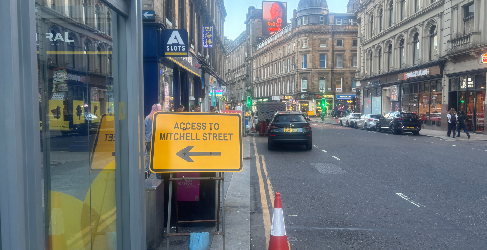 Drury Lane Diversion Urban street scene in Glasgow showing a yellow diversion sign reading 'Access to Mitchell Street' with a left-pointing arrow. The sign is placed on the pavement beside shopfronts, with cars parked along the road and pedestrians in the distance.