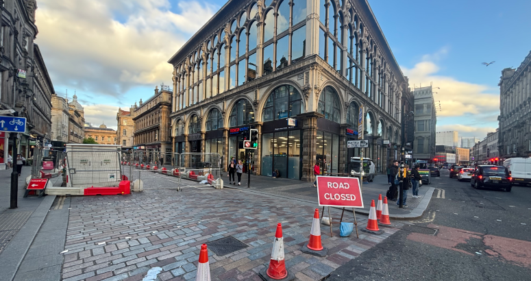 Gordon St, Glasgow, Road Closure A city street in Glasgow closed off with traffic cones and a 'Road Closed' sign due to sewer repair works. Construction barriers and signage are visible, with a historic glass-fronted building in the background under a partly cloudy sky.