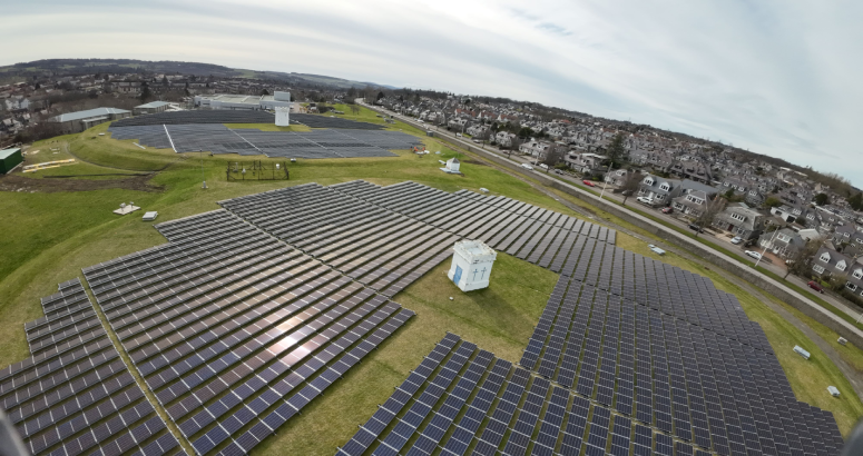 Drone image shows a circular formation of solar PV panels on grassed area, with houses in the background.