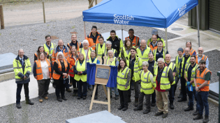 A large group of people wearing hi-viz PPE are standing outdoors, behind a wooden easel with stone plaque.