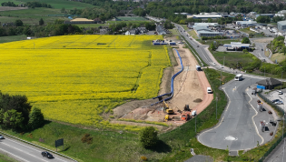 Newfield Project Progress Aerial view of a vibrant landscape showing a large bright yellow rapeseed field alongside roads with moving vehicles and construction equipment near a developing area. Rolling hills and greenery are visible in the background under a clear sky.