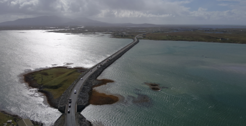 Aerial images taken of the South Ford Causeway Aerial view of a coastal road crossing over a narrow strip of land with shimmering blue waters on either side, leading towards distant mountains under a cloudy sky.