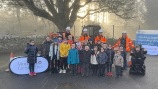 A group of children are standing outside in the school playground, facing the camera. There are four adults wearing orange hi-vis jackets behind them.