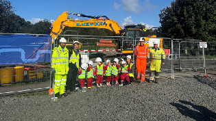 Children stand with mini high viz and hard hats on in front of a fence on a construction site. A yellow digger is behind them on the other side of the fence. The sky is blue and there are trees in the background.