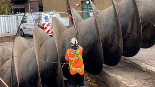 A person wearing hi-vis and PPE has their back to the camera; they're standing at the side of a 9 tonne metal screw being taken out of the ground