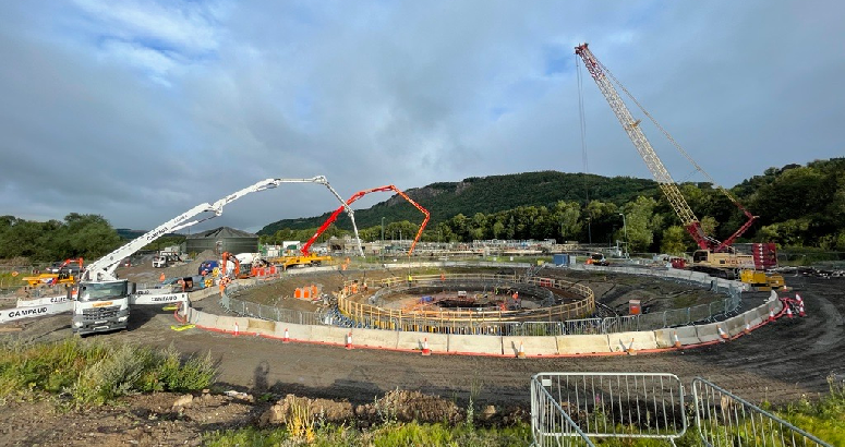 Construction site with multiple concrete pumps, a large crane, and workers engaged in building a large circular structure, set against a backdrop of green hills under a cloudy sky.