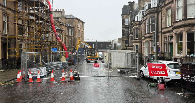 A city street is closed off for construction, with barricades, traffic cones, and a sign reading "Road Closed." Construction machinery is visible in the background, and scaffolding covers a nearby building. The weather is wet, with rain making the pavement glisten. Residential buildings line both sides of the street.