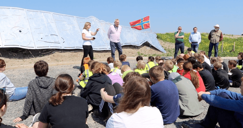 groups of children are sitting on the ground with their back to the camera. They are outside and facing some adults who are standing up in front of a metal sculpture of a whale.