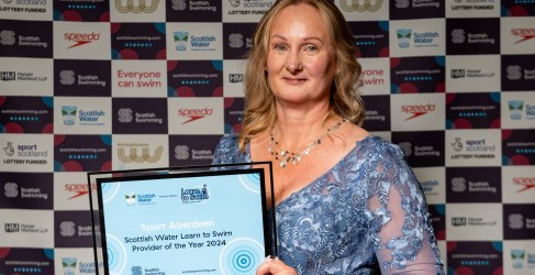 Person holding a "Sport Aberdeen Scottish Water Learn to Swim Provider of the Year 2024" award in front of a banner with various sponsors including Scottish Water and Speedo.
