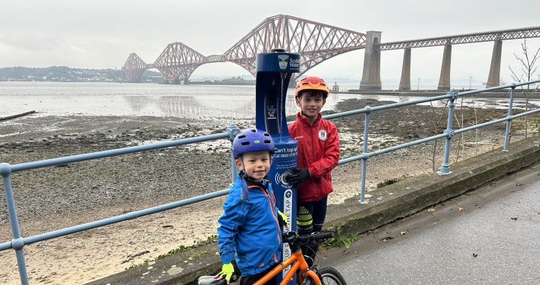 Two young children pose beside bright blue water refill fountain with several large bridges in the background