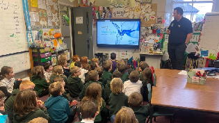 A group of young students in green uniforms sit on the floor of a colourful classroom, watching a presentation on a screen at the front. An adult stands nearby, delivering the talk. The room is filled with educational materials, posters, and decorations.