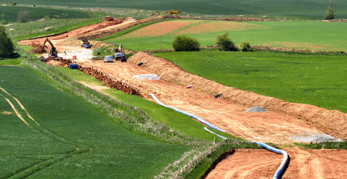 Construction site in a rural area with machinery working on a large trench surrounded by green fields.