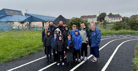 Pupils stand smiling with their headteacher and two employees of Scottish Water and one of George Leslie. They are standing on a running track, with buildings in the background.
