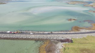 Aerial view of a causeway crossing a shallow, turquoise bay with scattered vehicles parked along the sides.