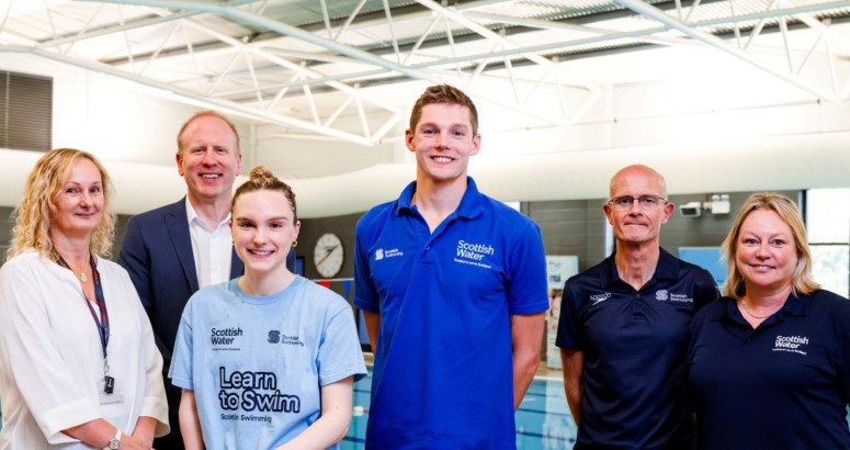 Group of six adults smiling in an indoor swimming pool facility; some wearing Scottish Water and Learn to Swim branded attire.