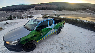 Scottish Water 4x4 vehicle beside a snowy reservoir dam with forested hills in the background.