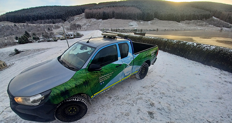 Scottish Water 4x4 vehicle beside a snowy reservoir dam with forested hills in the background.